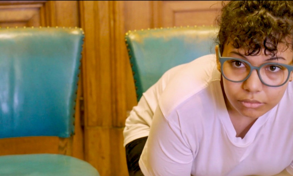 Film still from Reasonable Adjustments by Anna Macdonald. Woman wearing green glasses and a pink t-shirt, in the process of standing up or sitting down on green leather chair.