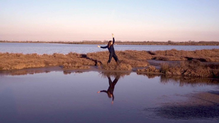 Film still from Intertidal. Barene by Laura Santini. Woman dancing on grassy piece of land, among still reflective water. 