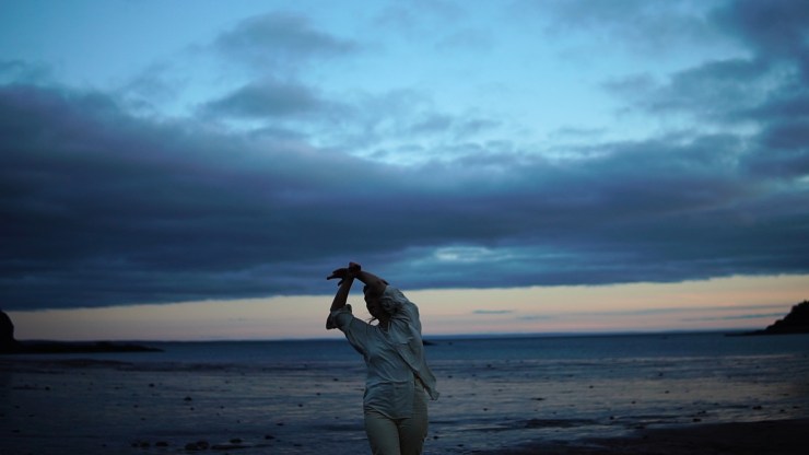 Film still from the moon rises in four parts by Michaela Gerussi & Tracy Valcarel. Person in motion in foreground of seaside landscape at dusk.
