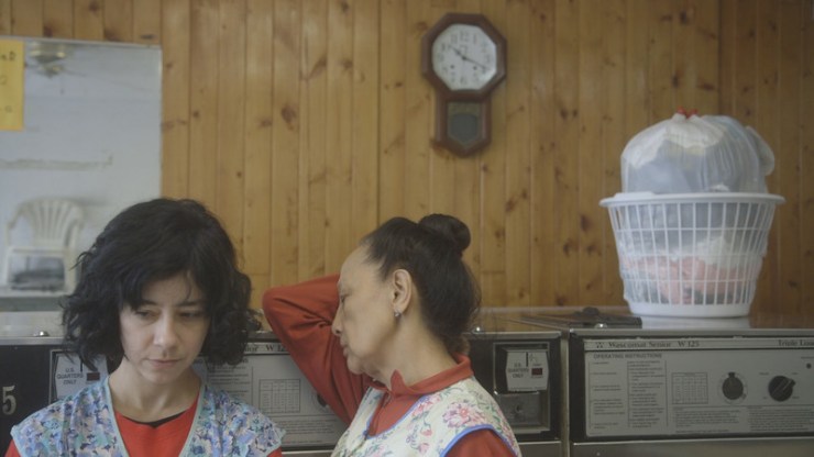 Film still from The Washing Society by Lynne Sachs & Lizzie Olesker. Two women rest in front of large washing machines, there is a clock on the wall and a laundry basket full of washing on top of the washing machines.
