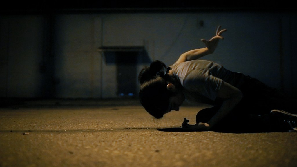 Film still from Unfurling by Alexa Velez. Woman kneeling on concrete ground, performing awkward hand gesture behind her back, in front of industrial looking building.