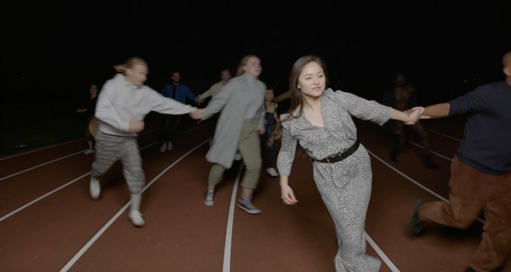 Film still from We Are Ready Now by Jack Thomson. People in motion, holding hands in pairs, on a marked out race track.